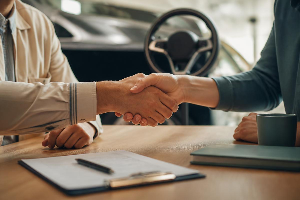 Duas pessoas se cumprimentando sobre uma mesa com um carro ao fundo